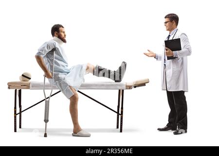 Verletzter Mann mit orthopädischem Stiefel und Halskragen, der auf einem Therapietisch sitzt und sich mit einem Arzt auf weißem Hintergrund unterhält Stockfoto