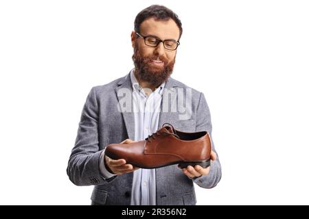 Ein junger bärtiger Mann, der einen eleganten braunen Lederschuh hält und isoliert auf weißem Hintergrund lächelt Stockfoto