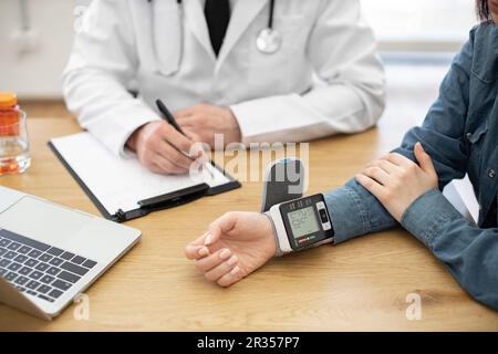 Schnittbild des weiblichen Handgelenks mit elektronischem Tonometer zur Darstellung des Blutdrucks. Der männliche Therapeut schreibt Zahlen auf Papier-Klemmbrett zur Regelung der Krankheitsbehandlung in der Klinik. Stockfoto