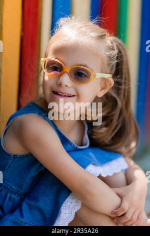 Ein kleines Mädchen mit Daunensyndrom, das eine Brille zum Sehen trägt, sitzt im Sommer neben einem farbigen Zaun in einer Kinderstadt. Hochwertiges Foto Stockfoto