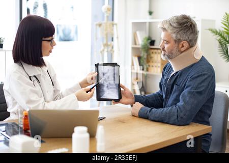 Seitenansicht einer jungen Frau in Brille und Labormantel, die auf eine Tablette zeigt, mit Röntgenstrahlen, die von einem attraktiven Mann mit Halsband gehalten werden. Hausarzt diskutiert Testergebnisse mit Patienten im Krankenhaus. Stockfoto