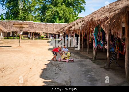 Zwei kleine Jungen spielen mit einem bunten Kinderfahrrad in einer ländlichen Umgebung in Thailand. Eine fröhliche Atmosphäre, die die Freude des Kindheitsspiels einfängt Stockfoto