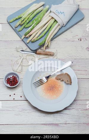 Weißer und grüner Spargel auf einem Holzbrett mit Schalen und Schäler, die Reste der Vinaigrette auf einem Teller mit einem Stück Brot und einer Gabel Stockfoto