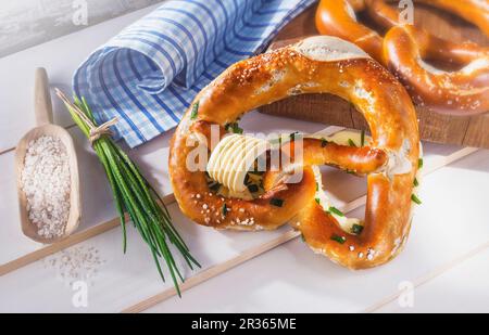 Laugenbrotbrezeln mit Butter und Schnittlauch neben einer Löffel Salz und einem Bündel Schnittlauch auf einer weißen Holzoberfläche Stockfoto