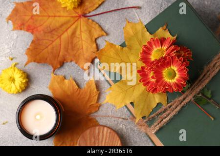 Buch dekoriert mit Chrysanthemen, Herbstblättern und Duftkerze auf einem hellgrauen strukturierten Tisch, flach liegend Stockfoto