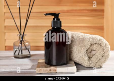 Eine Flasche Shampoo und ein gerolltes Handtuch auf einem weißen Tisch in der Nähe der Holzwand Stockfoto