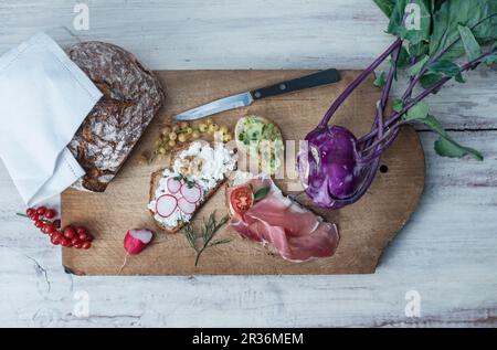 Scheiben Brot mit verschiedenen Toppings auf einem Schneidbrett mit Johannisbeeren, lila Kohlrabi und knusprigem Brot Stockfoto