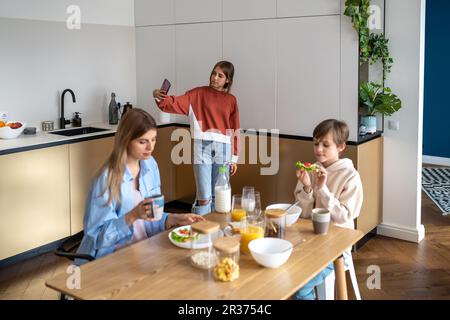 Familienvormittag, Tochter macht Selfie für soziale Medien am Telefon, mom und Sohn frühstücken. Stockfoto