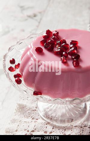 Rosafarbenes Milchgelee mit Granatapfelsamen auf einem Kuchenständer aus Glas und klassischer Tischdecke aus Spitze Stockfoto