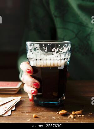 Ein Glas betrunkenen Guinness mit einer Damenhand mit roten Nägeln und einem grünen Pullover, der Glas an einem Holztisch hält, umgeben von Nüssen und Spielkarten Stockfoto