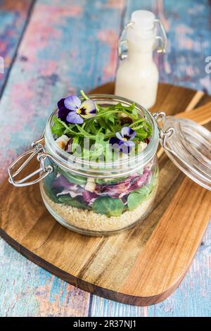 Ein quinoa Salat mit lämmer Eisbergsalat, Radicchio, Rucola, Croutons, Ziegenkäse und gehörnten Veilchen in einem Glas auf einem Holzbrett, mit dem Dressing in einer Stockfoto