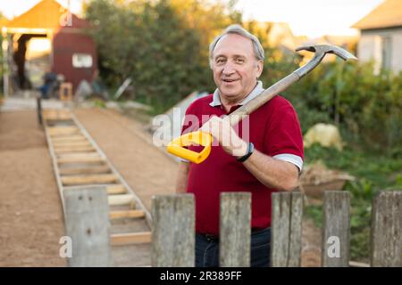 Ein erwachsener Mann stand entspannt neben dem Zaun, mit einer Schaufel im Garten, die lächelte Stockfoto