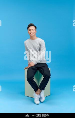 Blick auf den jungen Mann in Jeans und grauen T-Shirt-Sitzwürfel auf blauem Hintergrund Stockfoto