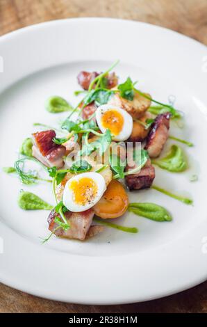 Gebratener Speck und Jakobsmuscheln in der Pfanne mit Wachteleiern und Erbsenpüree mit Erbsenschießen auf einem weißen Teller und einem Holztisch Stockfoto