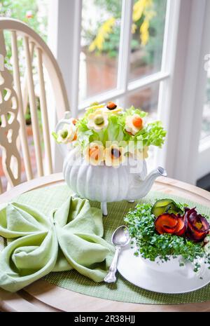 Strauß aus Salatblättern und Blumen mit Käse, Karotten und Avocado in einer Teekanne, Kresse und Karotten in einer Teetasse auf einem grünen Tablemat Stockfoto