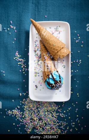 Blaues veganes Eis mit Schokoladensoße und Zuckerkonfetti in Zapfen Stockfoto