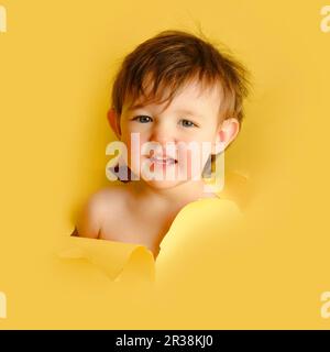 Trauriges Baby in einem Loch auf papiergelbem Hintergrund. Zerrissener Studiohintergrund des Kindes, Kopierraum. Ein Kind im Alter von einem Jahr, sechs Monaten Stockfoto