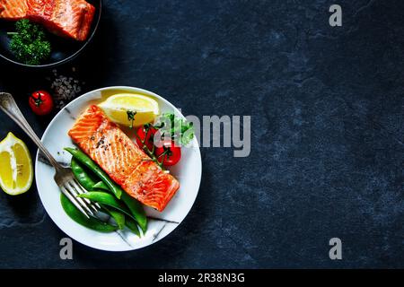 Gerösteter Lachs mit pochierten grünen Erbsen, Tomaten, Zitrone auf dem Teller und Lachsfilet in klassischer Bratpfanne Stockfoto