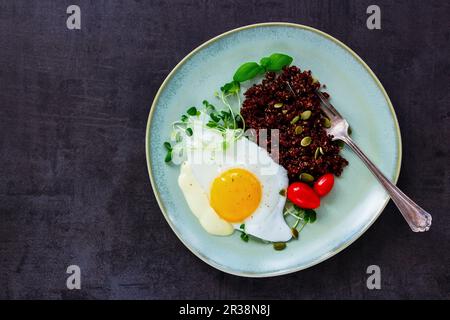 Teller aus roter Quinoa mit Spiegelei, Sprossen, Tomaten und Kürbiskernen für ein gesundes Abendessen auf dunklem, rustikalem Schieferhintergrund Stockfoto