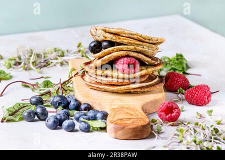 Stapel vegane Kichererbsen-Pfannkuchen auf Olivenholz Schneidebrett mit grünem Salat, Sprossen, Beeren, über grauem Küchentisch Stockfoto