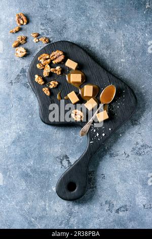 Gesalzene Karamellbonbons auf schwarzem Holzbrett mit Flour de sel, Karamellsauce und karamellisierten Walnüssen Stockfoto