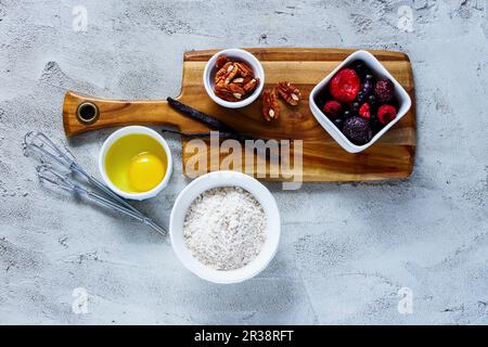 Holzschneidebrett mit Zutaten zum Backen (Vollmehl, Ei, gefrorene Beeren, Nüsse und Vanille) auf strukturiertem Betonhintergrund Stockfoto