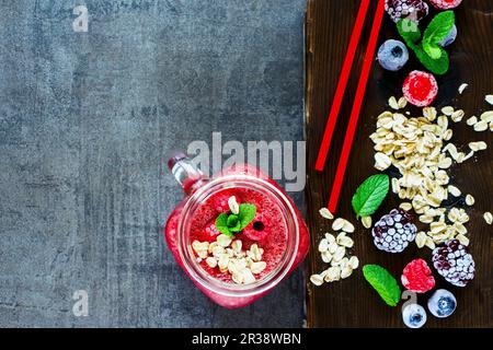 Nahaufnahme des roten Smoothie-Getränks im Glasgefäß und gesunde Zutaten auf dunklem Schneidebrett auf rustikalem Hintergrund Stockfoto