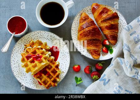 Nahaufnahme des Frühstückstisches mit frisch gebackenen Croissants und Waffeln, serviert mit Kaffee, Erdbeeren und hausgemachter Beerenmarmelade Stockfoto