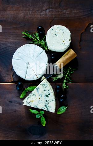 Blick von oben auf den rustikalen Küchentisch mit verschiedenen Käsesorten, Oliven, Basilikum und Rosmarin auf dunklem Holzhintergrund Stockfoto