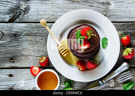Schokoladenpfannkuchen mit frischen Erdbeeren und Honig, serviert auf Keramikplatte auf altem grauen Holzhintergrund Stockfoto