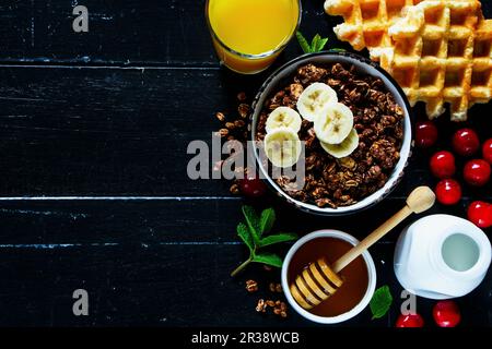 Schüssel mit Schokoladenhafergranola, Waffeln, Orangensaft, frischen Kirschen, Honig, Minze und Milch oder Joghurt Stockfoto