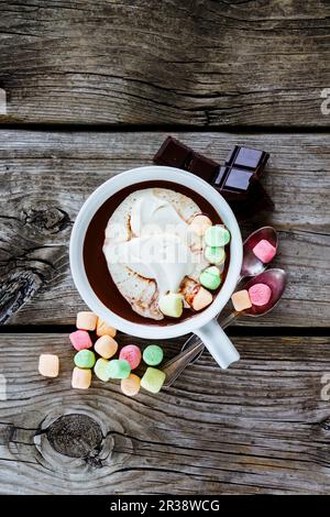 Nahaufnahme von heißer Schokolade in weißem Keramikbecher mit Schlagsahne und Marshmallows auf rustikalem Holzhintergrund Stockfoto