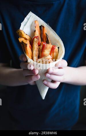 Ein Kind, das eine Mini-Schnitzel, Süßkartoffelfrites und Ketchup in einem Bambuskegel hält Stockfoto