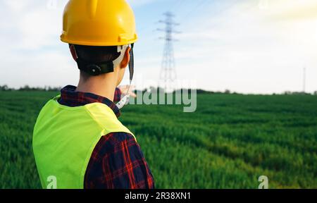 Bauarbeiter, der auf den Strompylon zeigt. Elektriker mit gelbem Schutzhelm und fluoreszierender Weste auf dem Feld, Arbeitszone. OSH. Stockfoto