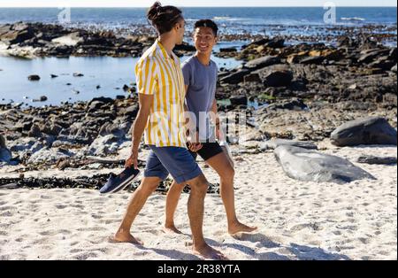 Glückliches birassisches, schwules Paar, das Händchen hält und barfuß am sonnigen Strand spaziert Stockfoto