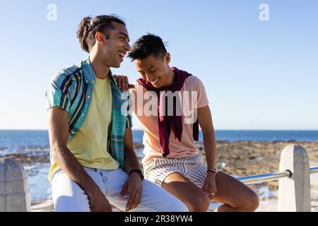 Ein glückliches, birassisches, schwules, männliches Paar, das auf der Promenade am Meer spricht und lächelt Stockfoto