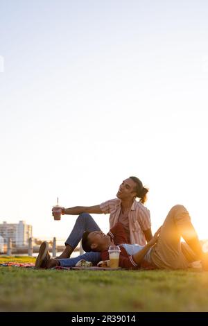 Glückliches, birassisches, schwules Paar, das bei Sonnenuntergang auf der Promenade am Meer picknickt, mit Kopierraum Stockfoto