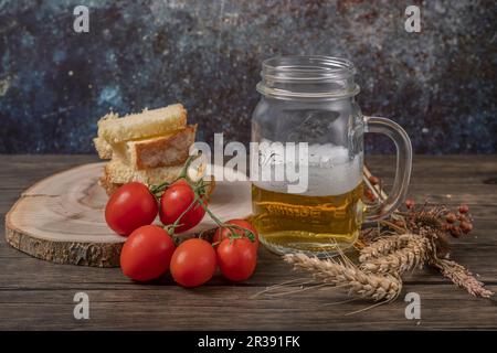 Eine Tasse Bier mit getoastetem Brot und roten Kirschtomaten daneben, mit getrockneten Maisohren Stockfoto