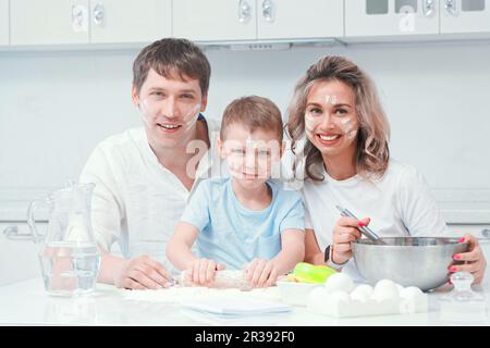 Eine wunderschöne, dreiköpfige, weiße Familie sitzt am Kochtisch, schaut in die Kamera und lächelt. Mit Mehl verschmutzt. Lustiges Foto. Stockfoto