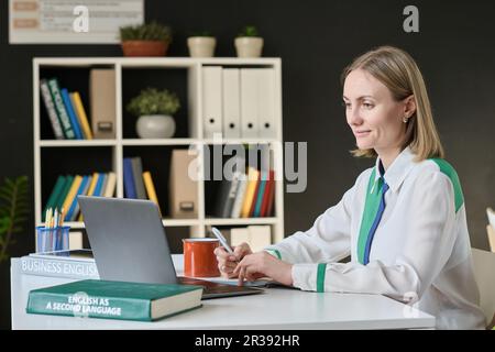 Junge Frau, die sich online auf den Unterricht vorbereitet, während sie mit einem Laptop im Unterricht am Tisch sitzt Stockfoto