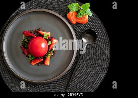 Draufsicht auf einen kugelförmigen Quark mit Erdbeeren und Brownies. Dessert mit glatten Oberflächen und Spiegelglasur auf dem schwarzen Teller. Schwarzer Hintergrund. Stockfoto