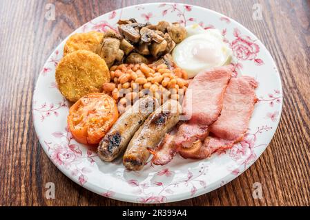 Traditionelles englisches Frühstück mit Speck, Würstchen, gegrillten Tomaten und Pilzen, gebackenen Bohnen, Kartoffelrösti Kuchen und Ei auf einem Holztisch Stockfoto