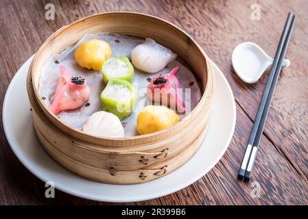 Bunte Dim Sum Knödel in einem Bambusdampfer auf einem Holztisch mit Essstäbchen Stockfoto