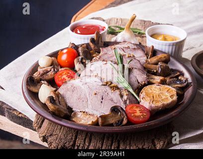 Scheiben gebackenes geröstetes Fleisch. Kalt gekochtes Schweinefleisch mit Pilzen, Knoblauch und Kirschtomaten auf braunem Teller Stockfoto