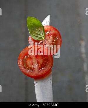 Tomatenscheiben mit einem Basilikumblatt auf einer Messerspitze mit Wassertröpfchen Stockfoto