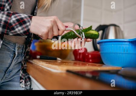 Weibliche Person, die eine Gurkenhaut mit einem Schälwerkzeug abzieht. Hausfrau bereitet frischen vegetarischen Salat mit kohlenhydratarmem Gemüse in einer Hausküche zu Stockfoto