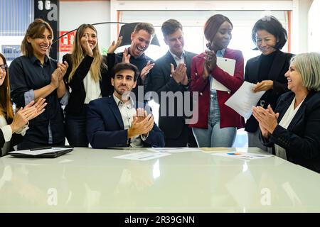 Eine Gruppe fröhlicher und vielseitiger Kollegen in formeller Kleidung, die im Büro am Tisch stehen und applaudieren, während sie ihren Erfolg feiern Stockfoto