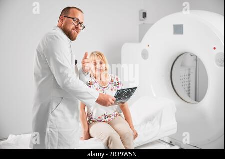 Medizinische Computertomographie oder MRT-Scanner. Arzt, der die MRT-Ergebnisse hält und Daumen nach oben zeigt. Patientin nach MRT, sitzend auf dem CT-Scan-Bett. Konzept der Medizin, des Gesundheitswesens, der modernen Diagnostik. Stockfoto