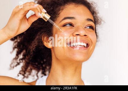 Süße lächelnde junge afrikanische Frau trägt feuchtigkeitsspendendes Serum aus der Pipette auf ihre Wange auf. Reizende, charmante Mischrasse, die sich um ihre Gesichtshaut kümmert. Stockfoto