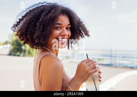 Glückliches afroamerikanisches Teenager-Mädchen, das Limonade im Open-Air-Café trinkt. Lächelnde, dunkelhäutige Teenagerin, die mit einem gefrorenen Cocktail in der Hand läuft. Charmin Stockfoto